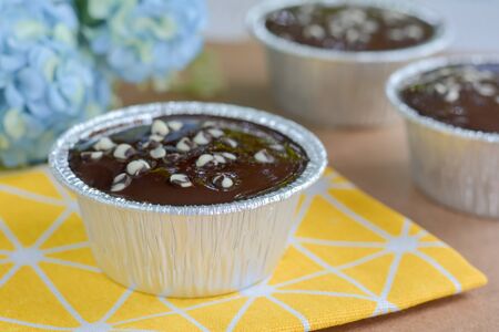 Dark chocolate cake with chocolate chips topping in round foil cup on yellow tablecloth and wooden table. Copy space for your text. Selective focus.の写真素材