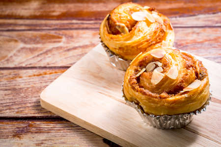 Delicious homemade almond danish bread in foil cup on a wooden chopping board and table in the home kitchen on black background.の写真素材