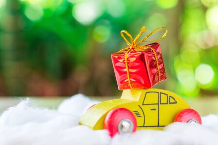 Red giftbox on roof of yellow wooden car on white cotton with green bokeh background, copy space for your text. Christmas and new year concept.の写真素材