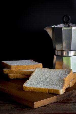 Homemade bread slides on a wooden chopping board in the kitchen in the house on a black background and aluminum Moka pot coffee maker and copy space. High carbohydrate food concept.の写真素材