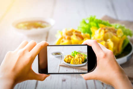 Food photographer and social media concept. Hand holding black smartphone and take dumpling or dim sum photo for share on blog, website, and social media channel.の写真素材