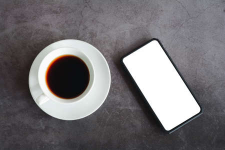 Business concept. Black coffee or hot espresso in a white ceramic cup and hand hold white screen on a smartphone or mobile on a dark table.の写真素材