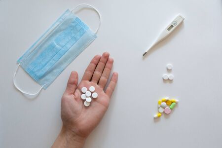 In the hands of the doctor is an electronic thermometer for measuring temperature, a protective mask from coronavirus and tablets, medications on a blue background .COVID-2019の写真素材