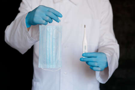 Doctors hand in a rubber glove holding,digital clinical thermometer with a high temperature indication of a sick patient and medical face mask against a black backgroundの写真素材