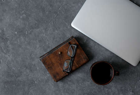 Brown leather notebook, pen, laptop and glasses on gray background.