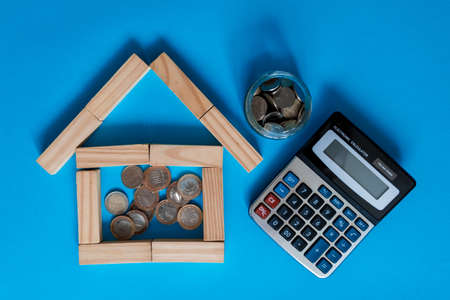 hand inserting coin in wooden model house over coin stack on green fake grass and calculator with...