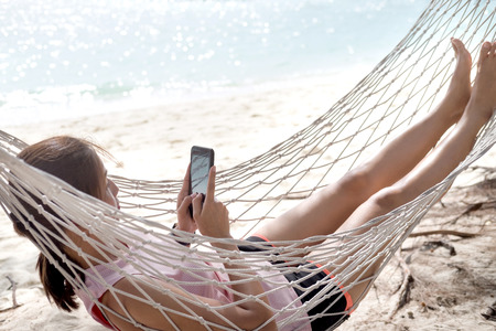 Woman using mobile phone on cozy mesh hammock at sea beach. Relaxation vacation and chilling on the sea beach.の写真素材