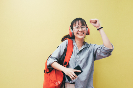 Teen lifestyle concept. Happy pretty hipster girl in earphones listening to music and dancing and holding mobile phone on bright yellow background.の写真素材
