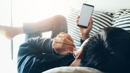 A young man lying down on a soft cushion feel relaxed and listen to online music from mobile phone...