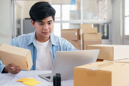 Young business startup online seller owner using computer for checking the customer orders from email or website and preparing packages for product office equipment.の写真素材