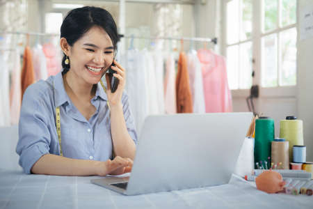 Young tailor design owner woman talking with customer on smart phone in her studio with clothes hanging in the background. Business owner shop and entrepreneur concept.の写真素材