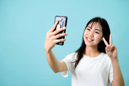 Young brunette in white shirt showing two fingers and taking selfie with smartphone on blue...