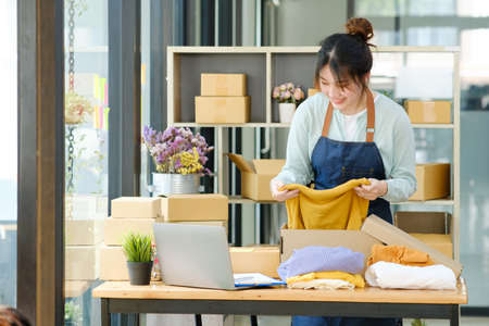 Asian female clothes shop owner folding a t-shirt and packing in a cardboard parcel box.
