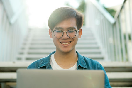 Young male college student with eyeglasses and in casual clothings smiling and sitting outdoor on stairs to study using laptop looking for information for school project and research. Education and E-Learning concept.の写真素材
