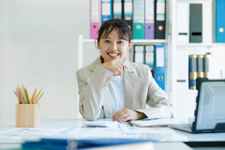 Yong Asian confident businesswoman is smiling and looking at camera sitting at working desktop in modern office. Business online worker concept.の写真素材