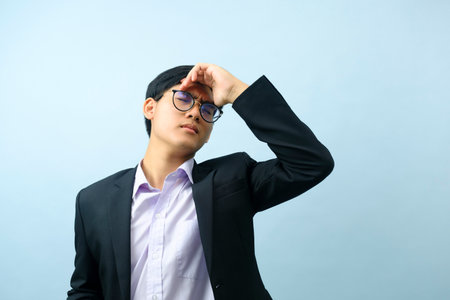 Portrait of young determined asian businessman wearing eyeglasses standing and putting his hand on his head stressed and frustrated from business work with light blue isolated background. Business concept.の写真素材