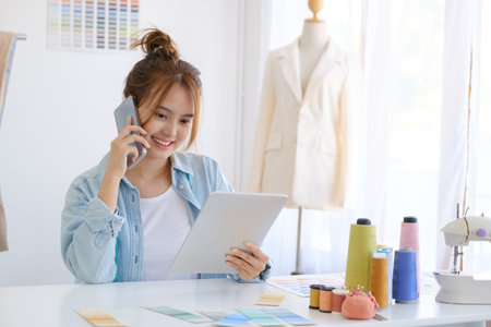 Young asian professional fashion designer is a working woman. She is sitting and talking on mobile consulting client about her suit and about online order in fashion. She also give a consultation for her order in the fashion shop.の写真素材