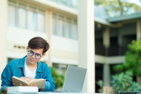 Young male college student with eyeglasses and in casual clothings sitting outdoor to study, read and holding book using laptop searching for information for school project and research. Education and E-Learning concept.の写真素材