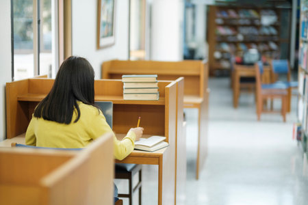 Young female college highschool student wearing casual clothings sitting at private desk reading book, studying, writing, and doing research for school project at a library. Learning Educational Library concept.の写真素材