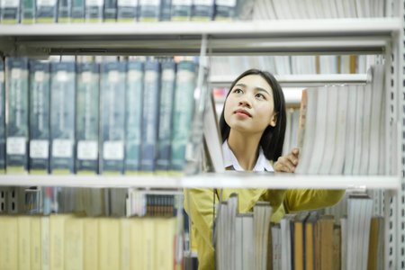 Young asian female student in casual outfit standing, looking and choosing books to read at library for studying, academic research, or school work or project. Learning, Education, Library concept.の写真素材