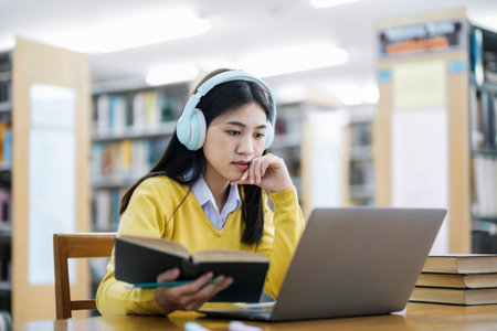 Young female college, highschool student wearing headphones and in casual clothings sitting at desk reading book, studying and doing research using laptop for school project at a library. E-Learning and Educational concept.の写真素材