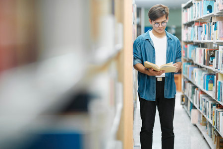 Young male student in casual outfit with backpack standing, reading or choosing books to read at...
