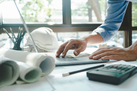 Close-up hands shot of engineer, developer, or architect standing and working alone at desk with papers, blueprints, pencils, calculator, and helmet using laptop for design or construction work. Engineer concept.の写真素材
