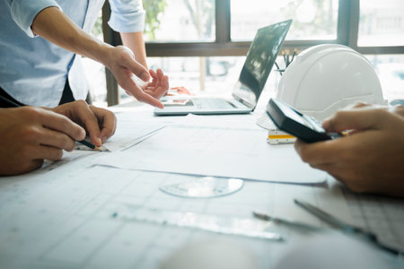 Close-up hands shot of three engineer, developers, or architects working together, drawing graphic design of construction project, and discussing for improvement at desk with papers, compass, helmet, calculator, rulers, and laptop. Engineering and Teamwork concept.の写真素材