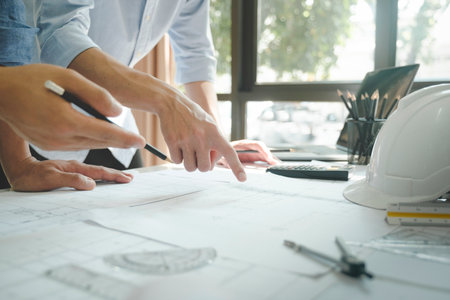 Close-up hands shot of two engineer, developers, or architects working together, drawing graphic design of construction project, and discussing for improvement at desk with papers, compass, helmet, calculator, rulers, and laptop . Engineering and Teamwork concept.の写真素材