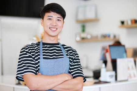 Cheerful young Asian man entrepreneur standing at counter in his own coffee shopの写真素材
