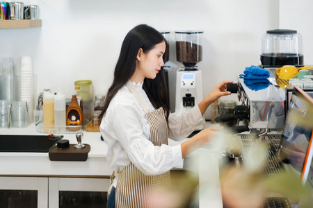 Young cheerful barista wearing apron while preparing coffee at an automatic machine in a modern coffee shop.の写真素材