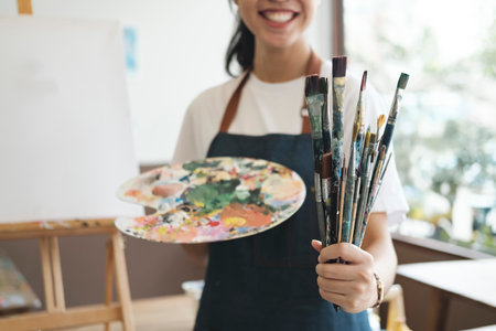 A close-up of the artist hands wearing an apron smeared with paint. Clutching many brushes and...
