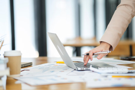 Close-up of a businesswoman hand as she meticulously analyzes data on her laptop, surrounded by business charts and a calculator.の写真素材