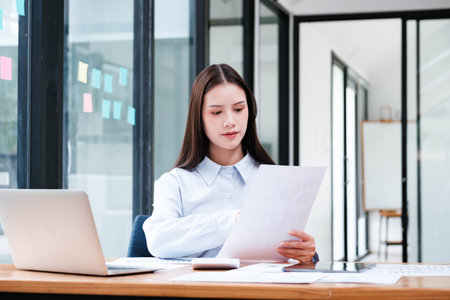 A focused female professional intently reviewing a document at her office desk with a laptop and smartphone nearby.の写真素材