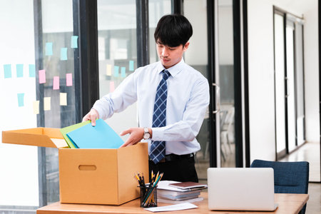 A man in a suit is opening a cardboard box on a desk. The box contains papers and a laptopの写真素材