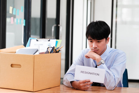 Contemplative Man at Office Desk with Packed Box and Resignation Letter, Reflecting on Career Change and Next Stepsの写真素材