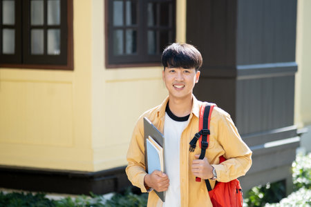 A happy and content young man stands outside on his university campus, holding a laptop and books, embodying the optimism of college life.の写真素材