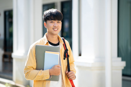 A contented college student carrying a laptop and books walks across the campus grounds, exuding a sense of readiness and enthusiasm for learning.の写真素材