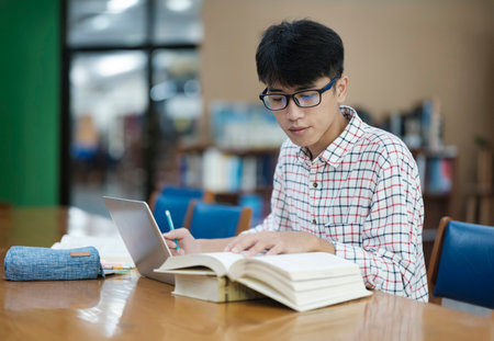 Young Asian male sitting inside a library alone doing research. Man working on a project. Young man doing research for a case.の写真素材
