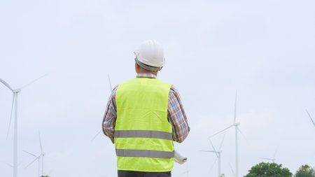 Engineer holding blueprint while surveying wind turbines at renewable energy site under bright sky conditions.の写真素材