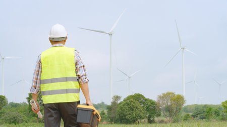 Engineer walking through wind farm with toolbox and blueprint for renewable energy maintenance and inspectionの写真素材
