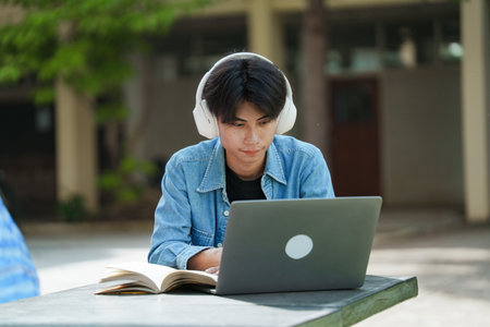 Portrait of a young Asian male college student e-learning using laptop on the bench on campus.の写真素材