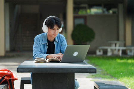 Portrait of a young Asian male college student e-learning using laptop on the bench on campus.の写真素材