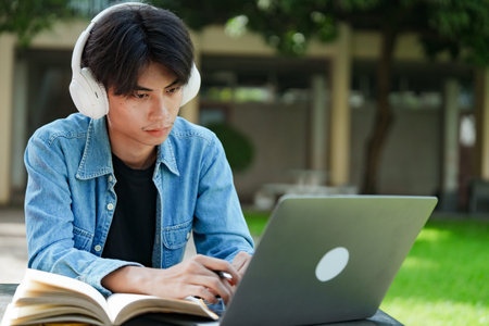 Portrait of a young Asian male college student e-learning using laptop on the bench on campus.の写真素材