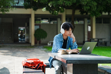 Portrait of a young Asian male college student e-learning using laptop on the bench on campus.の写真素材