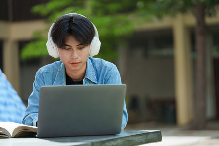 Portrait of a young Asian male college student e-learning using laptop on the bench on campus.の写真素材