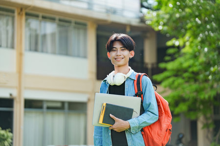A cheerful Asian college student stands outside on campus with a backpack, smiling and looking at the camera.の写真素材