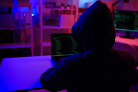A mysterious hooded hacker working on a computer in a dark room.の写真素材