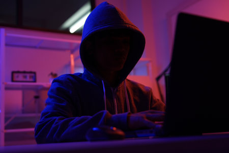 A mysterious hooded hacker working on a computer in a dark room.の写真素材