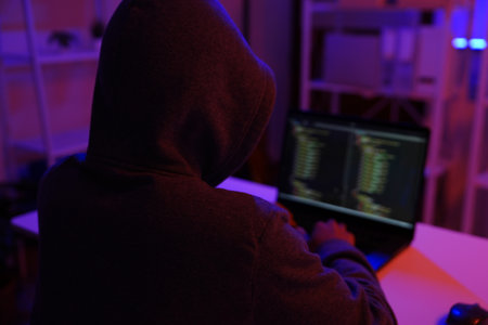 A mysterious hooded hacker working on a computer in a dark room.の写真素材
